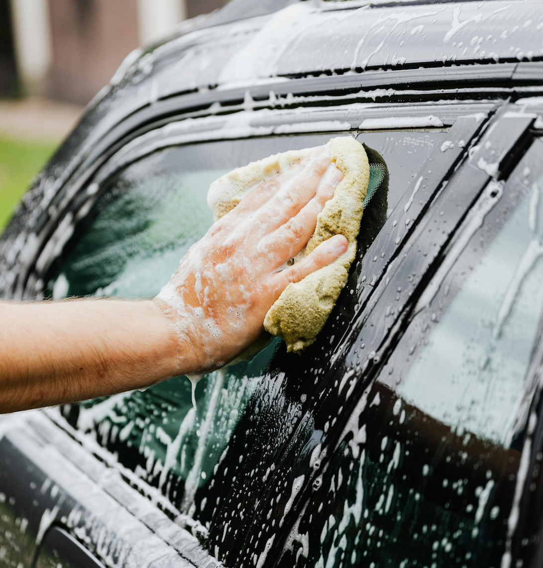 A person is washing a car outdoors, focusing on soapy water and sponge cleaning, highlighting the car's gleaming surface.